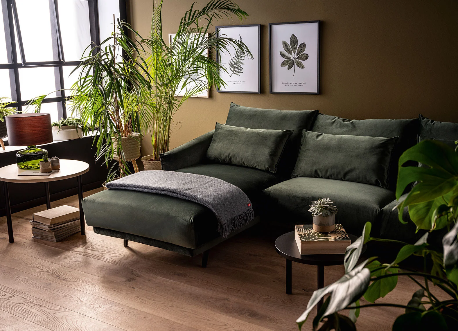Zielony narożnik w salonie z licznymi roślinami doniczkowymi i botanicznymi grafikami na ścianie.